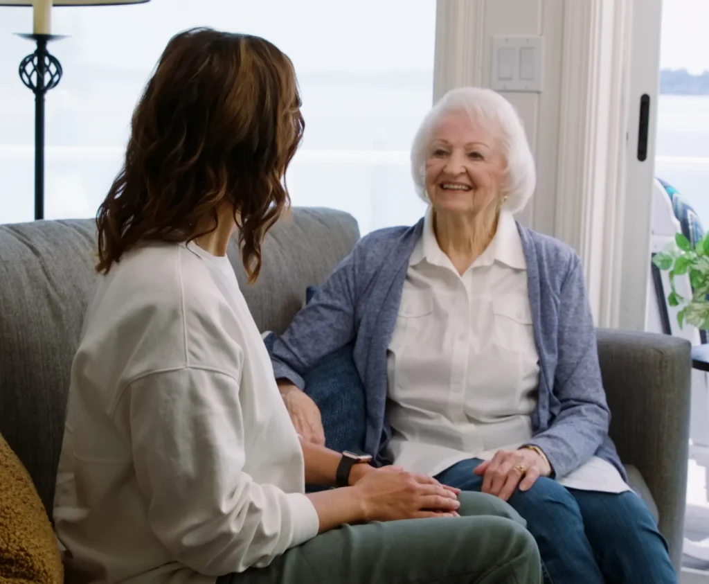 Smiling elderly woman sitting on a couch talking with a younger woman in a bright living room, promoting warm connections in senior living.